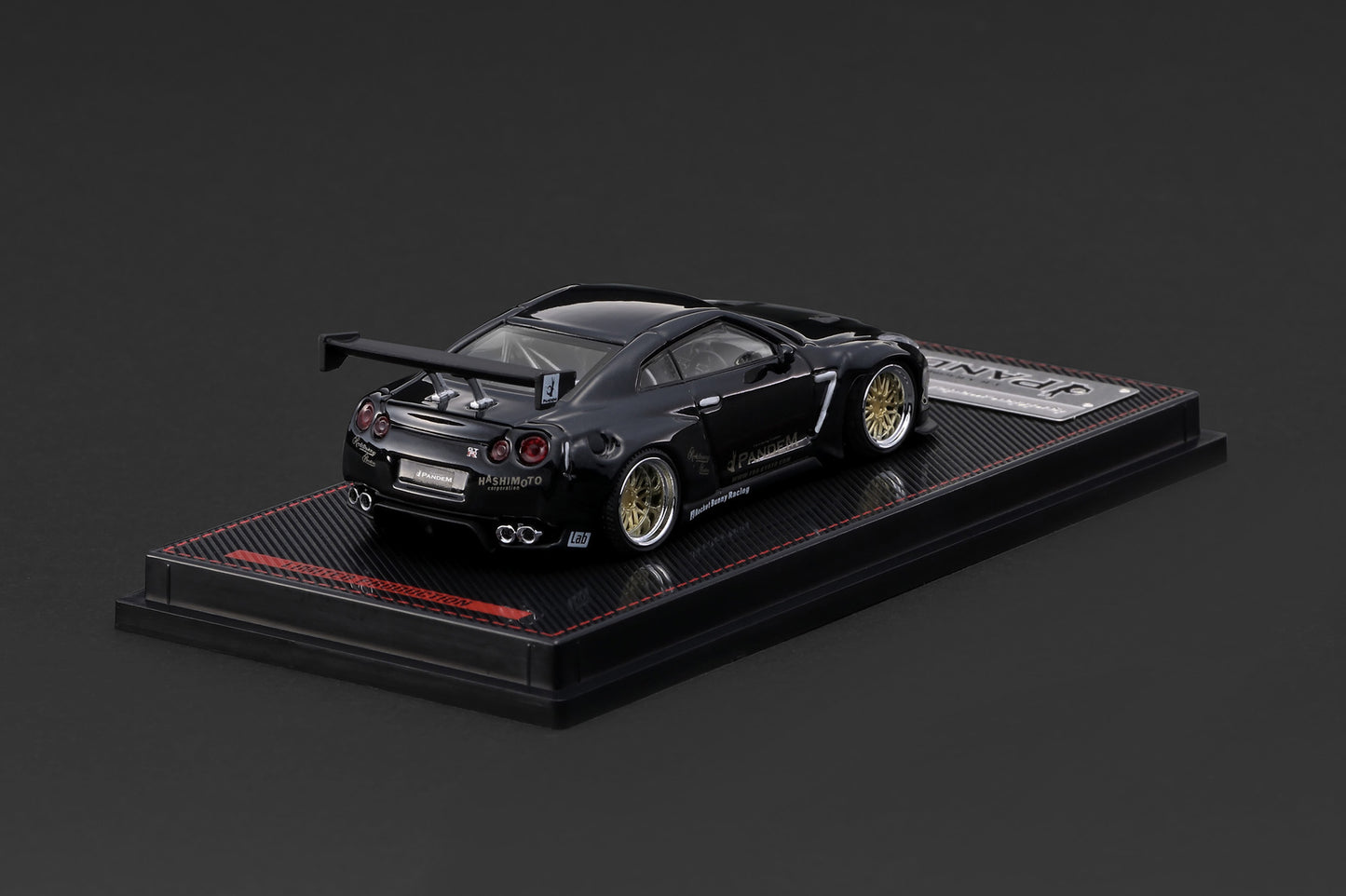 A black die-cast model of a Nissan GT-R Nismo race car, displayed on a black textured base with a small silver plaque, set against a dark gray background.
