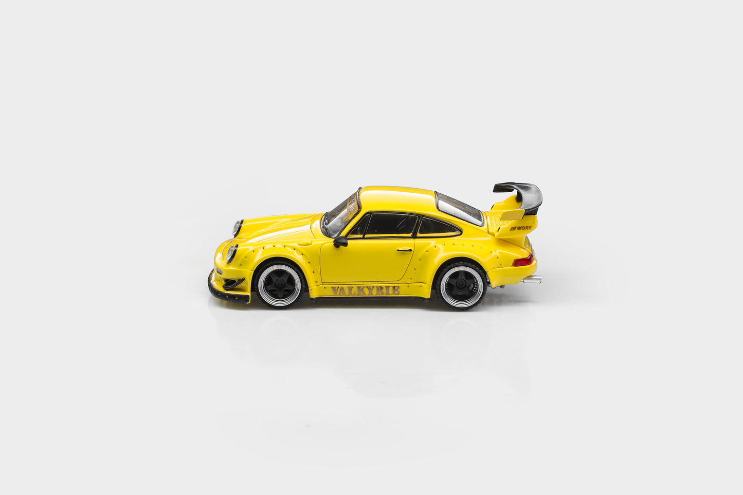 A yellow diecast model car resembling a Porsche 911 with a large rear wing and black wheels, shown from the side on a light gray background.