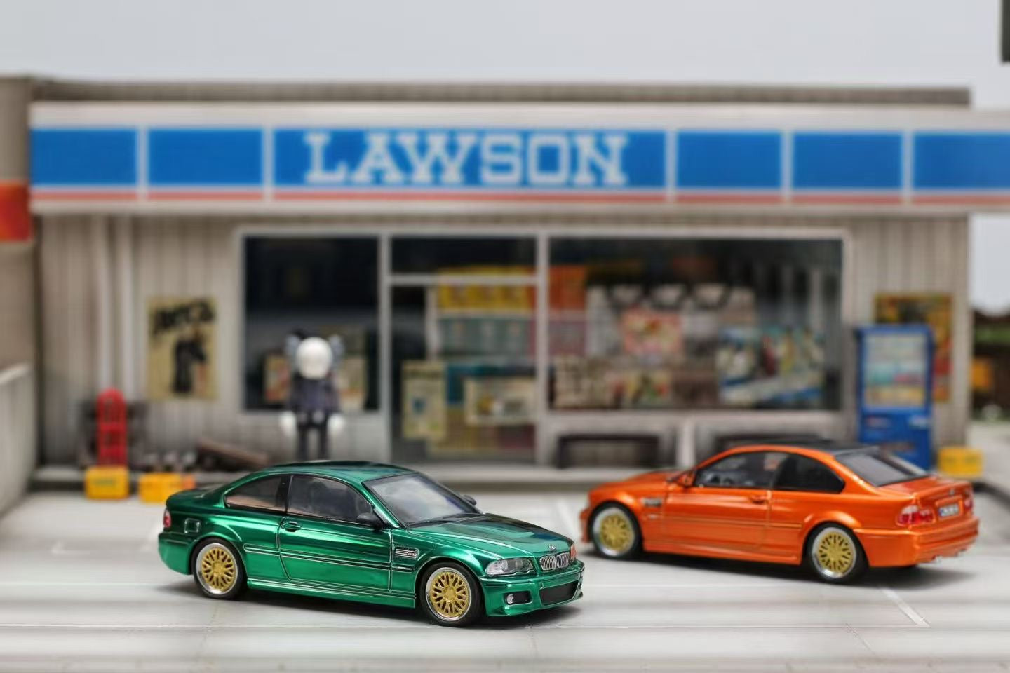 Two toy cars, one green and one orange, parked in front of a Lawson convenience store.