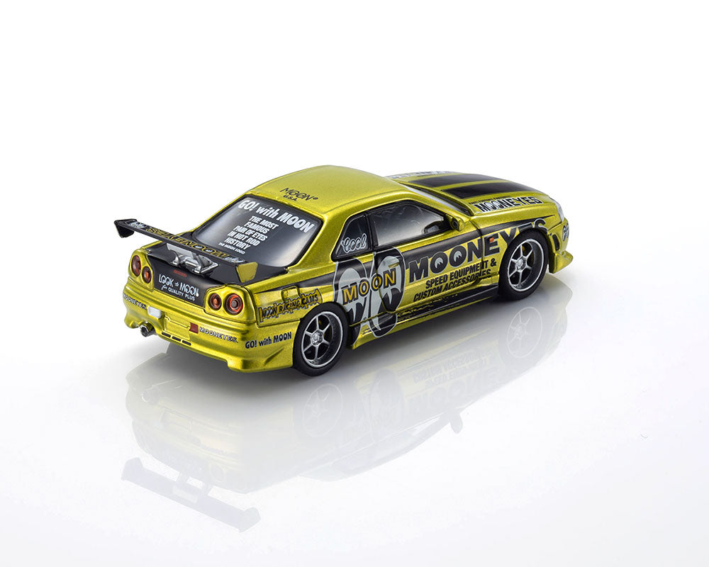 A metallic gold and black die-cast model car, a Nissan Skyline R34 GT-R, featuring 'Moon Moone' and 'Racing Team' decals, photographed on a reflective white surface against a white background.