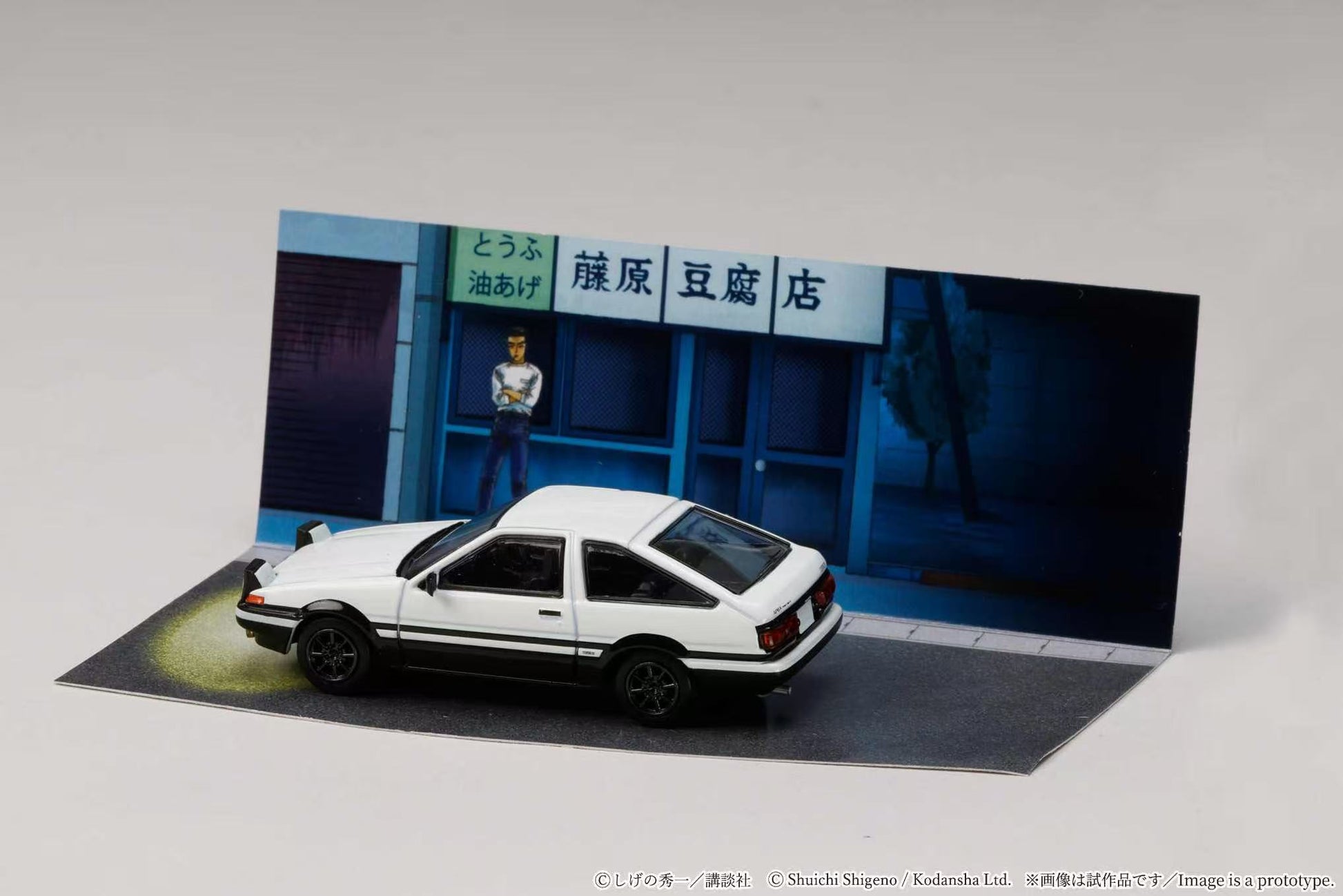 Model car in front of a miniature store with a person inside, on a gray background