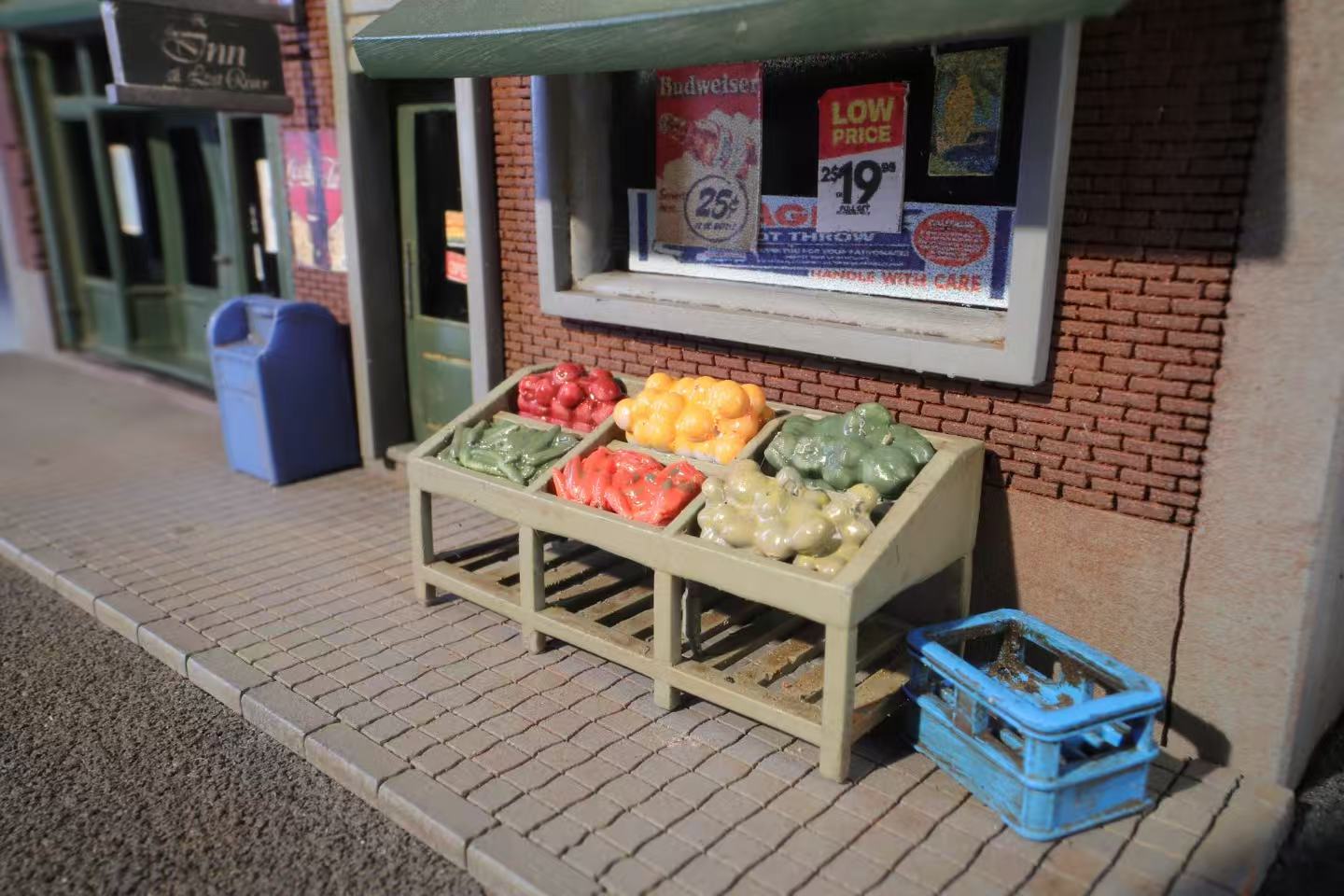 A miniature streetscape with a fruit stand displaying colorful produce: red apples, yellow oranges, green vegetables. Nearby, a blue crate and a vintage brick storefront with posters convey a nostalgic small-town feel.