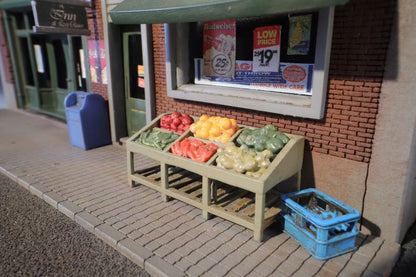 A miniature streetscape with a fruit stand displaying colorful produce: red apples, yellow oranges, green vegetables. Nearby, a blue crate and a vintage brick storefront with posters convey a nostalgic small-town feel.