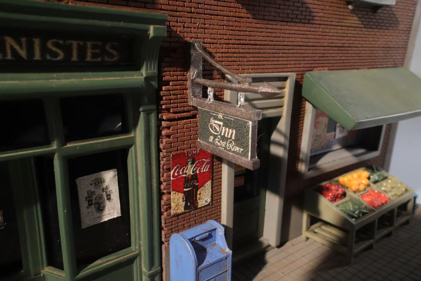 A quaint street scene with a vintage storefront. A sign reads "Inn & Sea River." Next to it, a blue mailbox and produce stand with colorful vegetables.