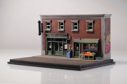 Miniature model of a brick corner store with green awning, fruit stand, and graffiti on the side wall. Evokes an urban, nostalgic feel.