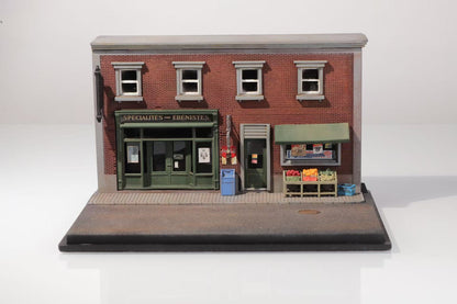 Miniature model of a vintage brick storefront labeled "Specialites des Ebenistes," with a green door, blue mailbox, and produce stand.