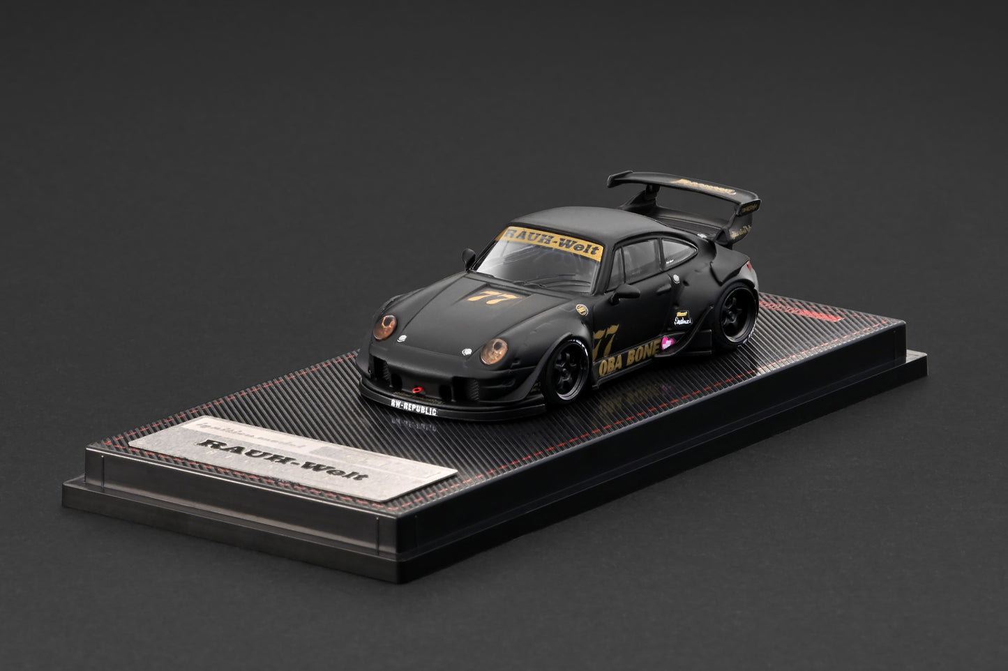 A black die-cast model car displayed on a branded black base against a dark gray studio background. The car has gold and white racing decals and the number 77.