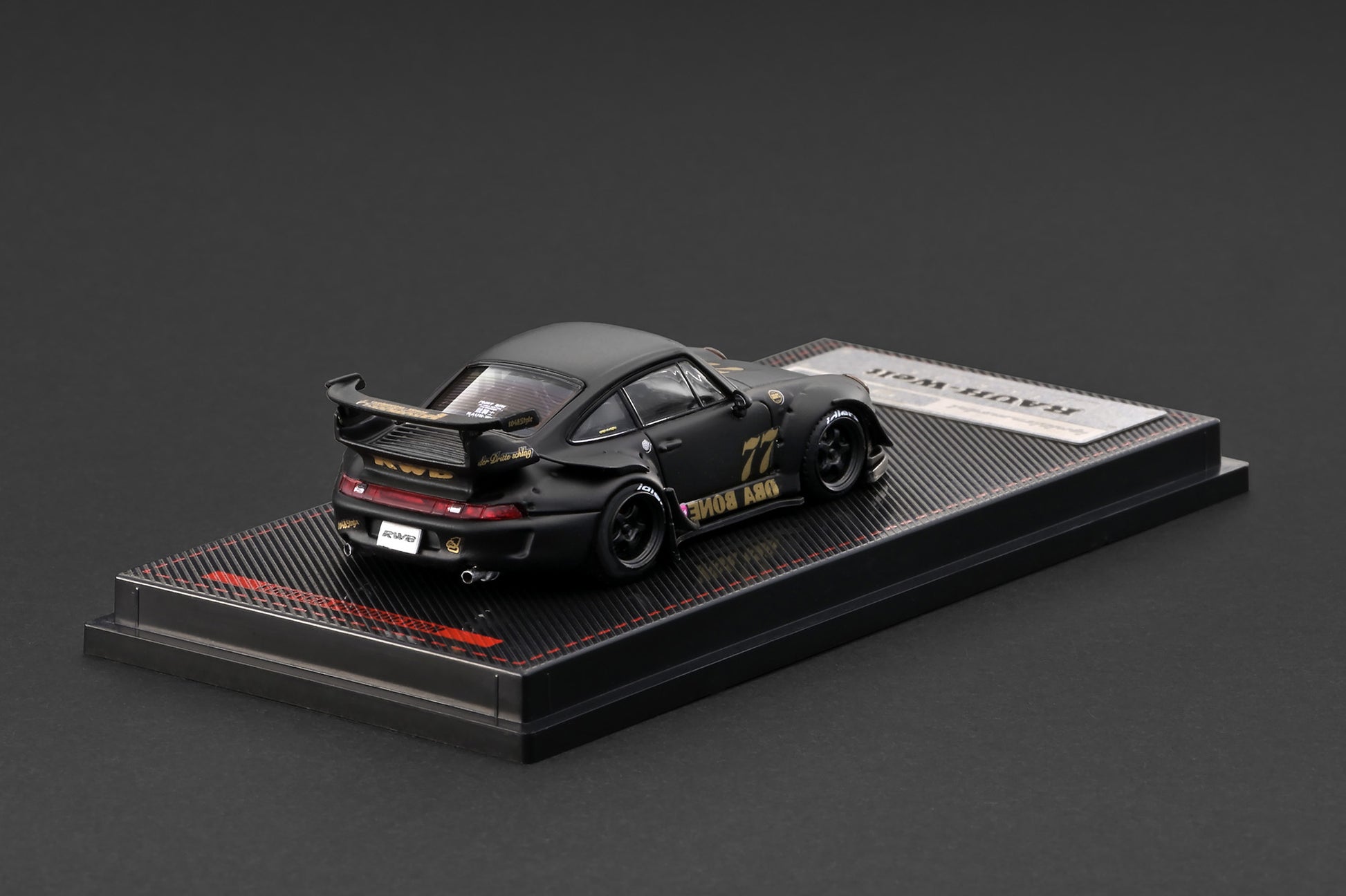 A black die-cast model car displayed on a branded black base against a dark gray studio background. The car has gold and white racing graphics, including the number 77 and 'TOSHIBA' branding, and is photographed at an angle showing its rear three-quarter view.
