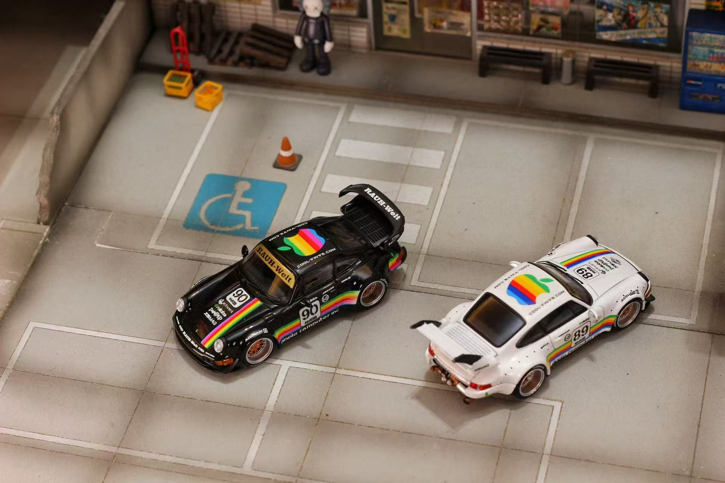 Two toy cars with rainbow designs on a miniature parking lot with accessibility symbol.