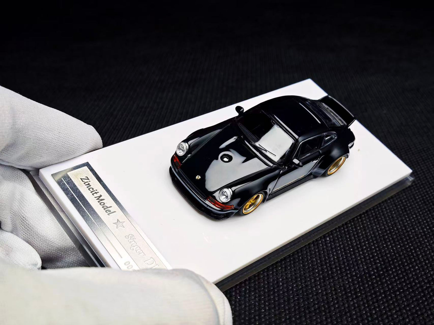 Black model car on a display stand with a white card, held by a gloved hand.