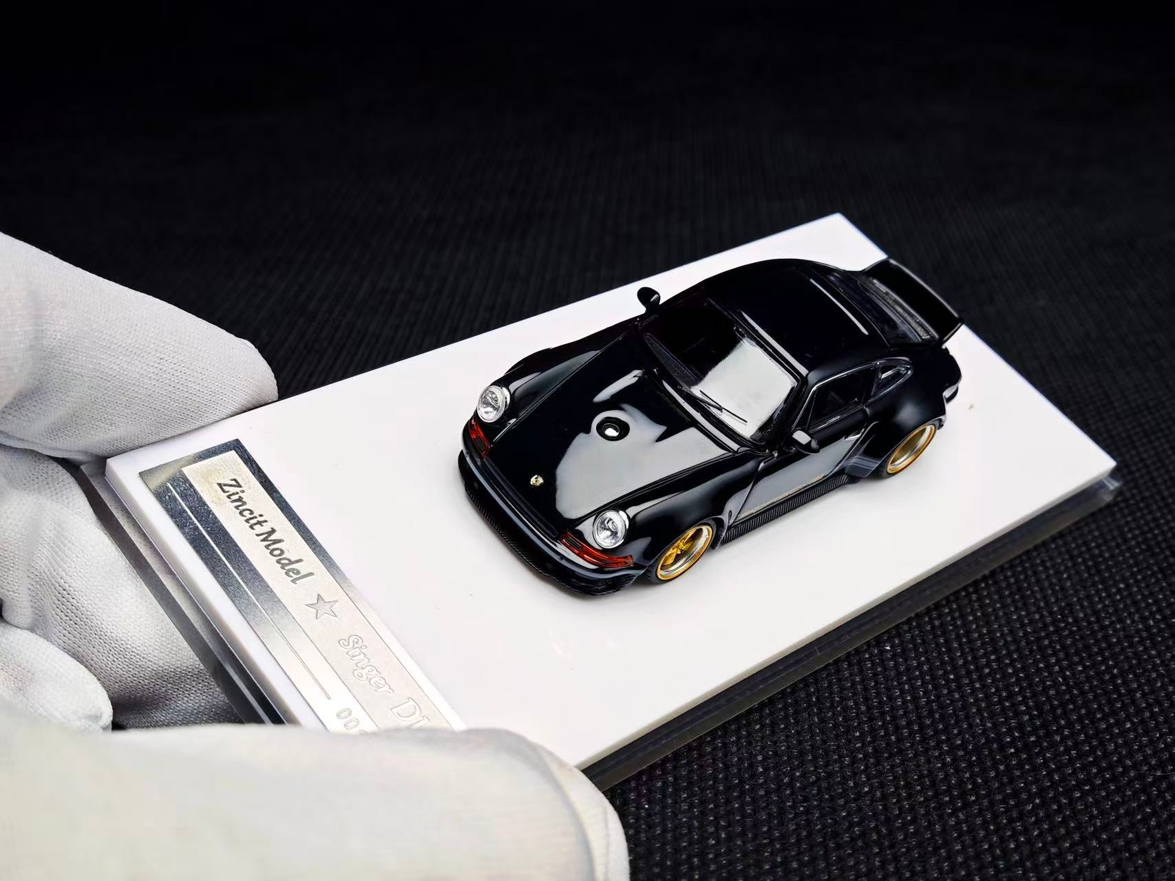 Black model car on a display stand with a white card, held by a gloved hand.