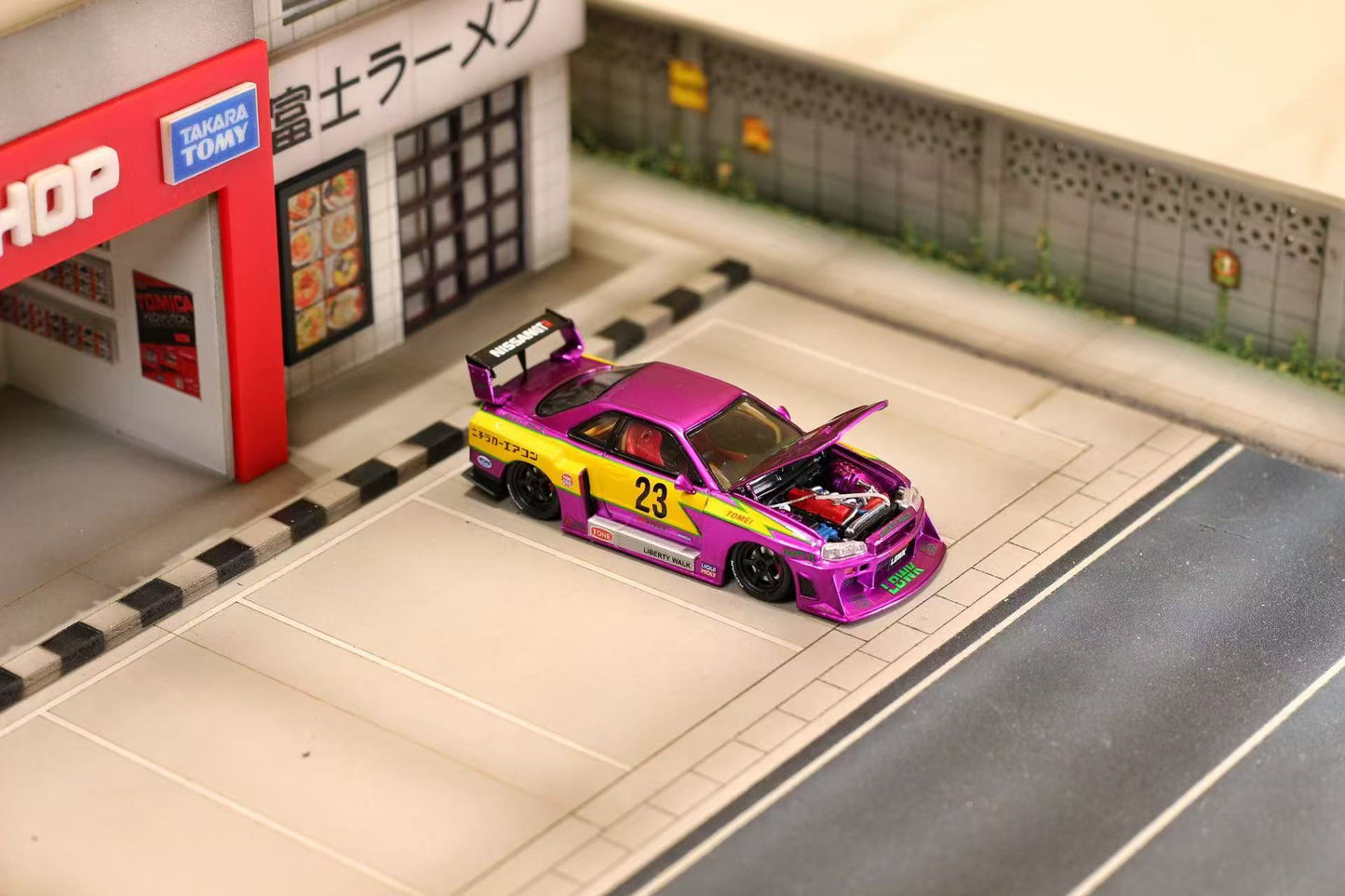 Model car with open hood on a miniature street scene in front of a store.