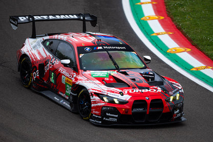 A red and white BMW M Motorsport race car with various sponsor logos, captured from a dynamic front-three-quarter angle on a racetrack with a green and white curbed section visible.