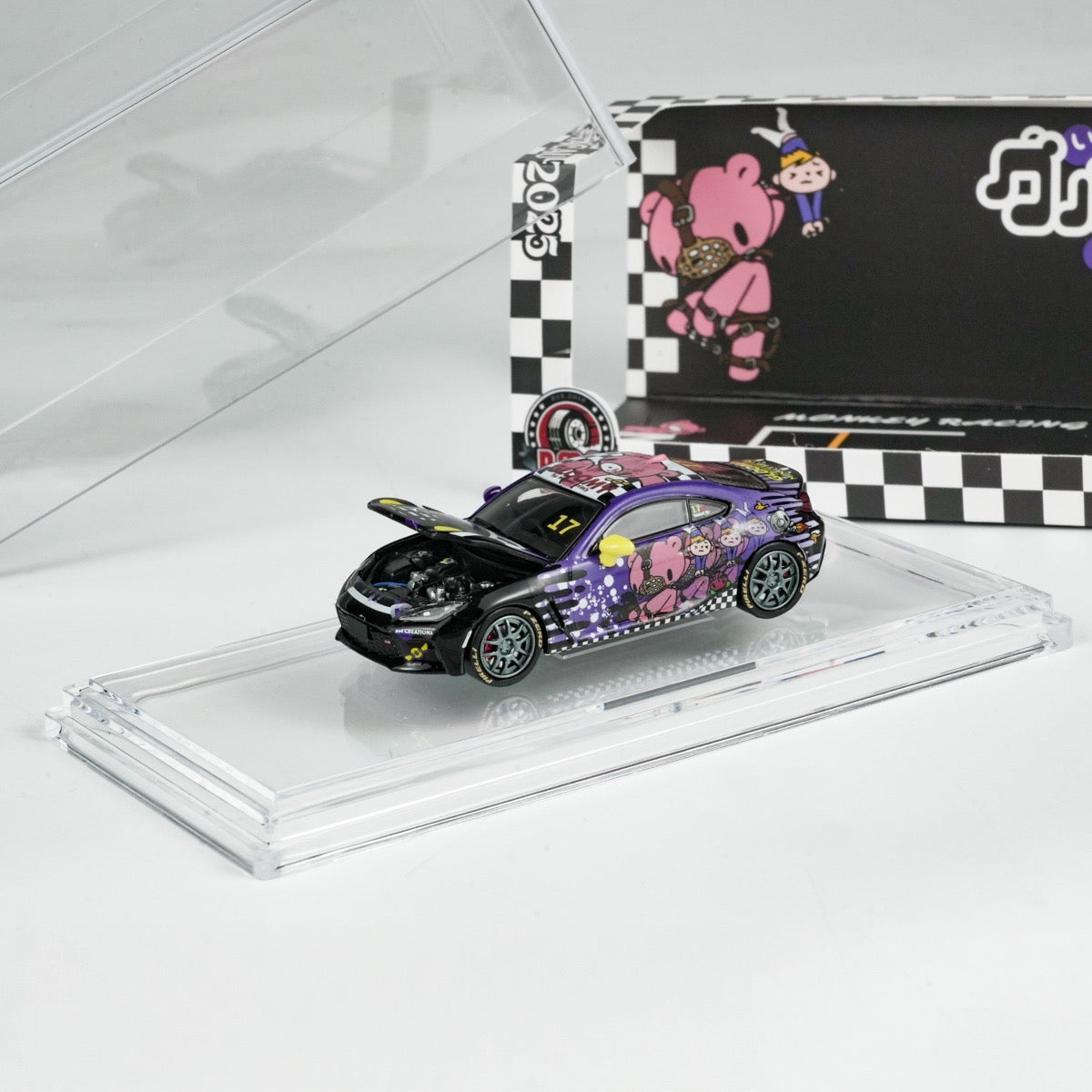 A toy car with a detailed engine and vibrant purple design featuring cartoon bears is displayed on a clear platform. Its box shows matching checkerboard patterns.