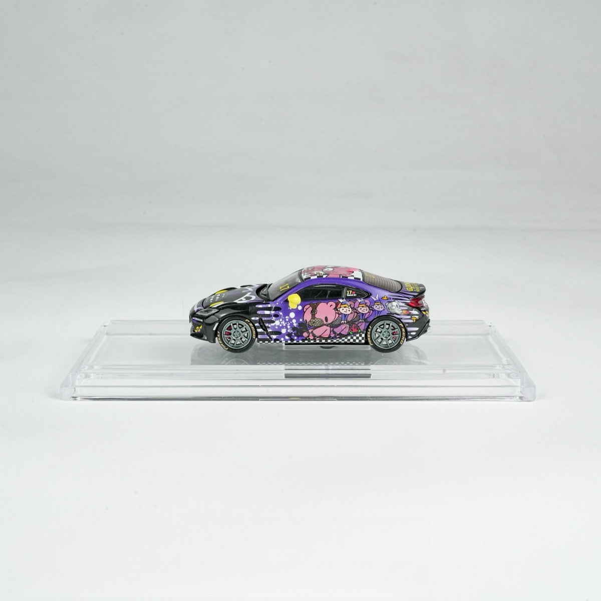 A toy car with vibrant graffiti art, including pink cartoon figures and black checkered patterns, sits on a clear display stand against a white background.