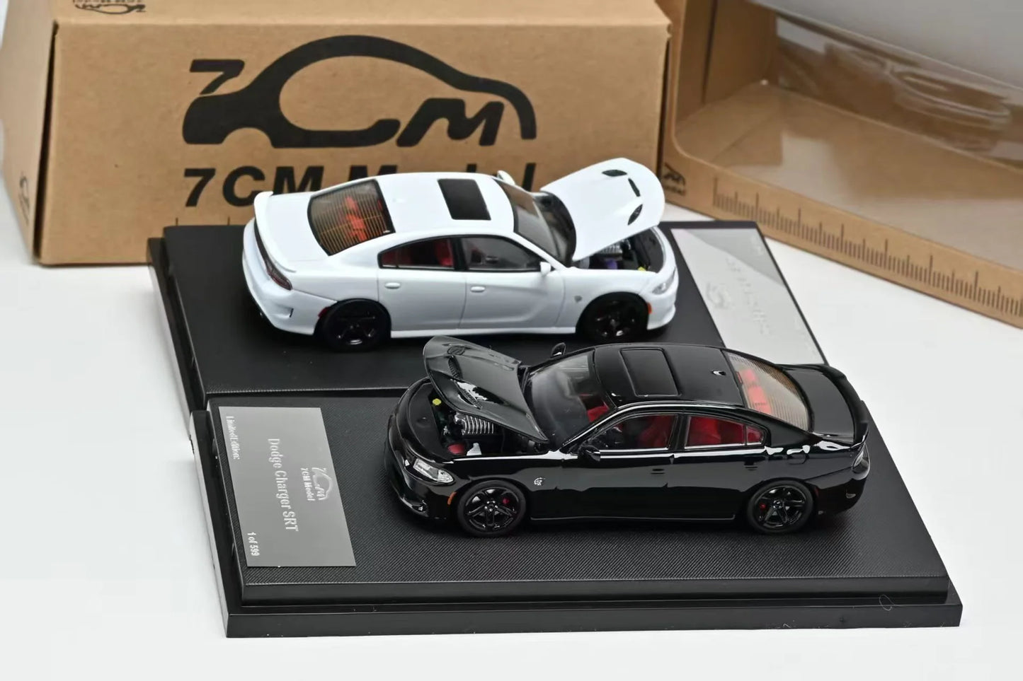 Two 1:64 scale diecast model cars from 7CM on black display stands, with a cardboard box in the background. The top car is white with its hood open, and the bottom car is black with its hood open.