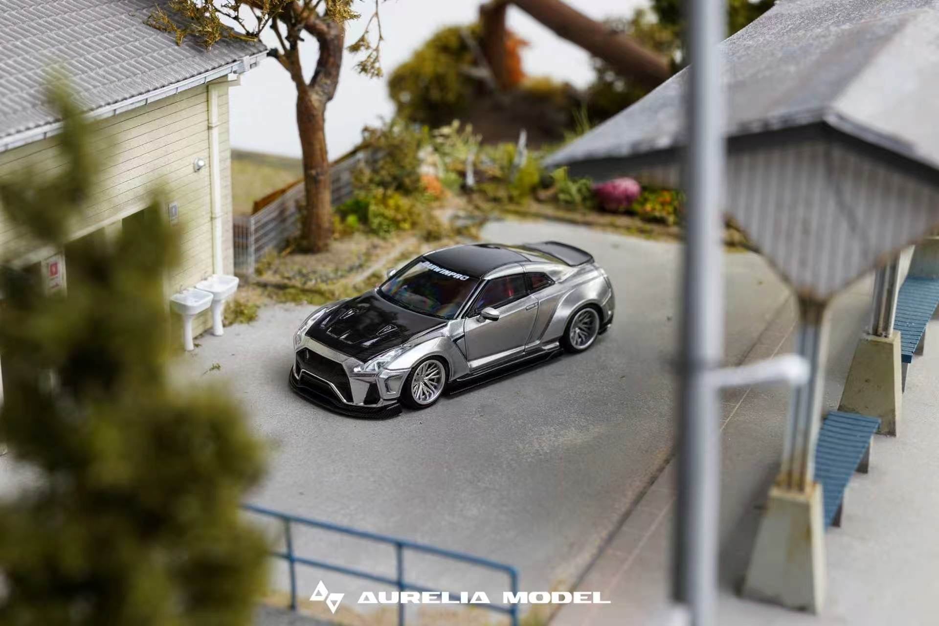 A miniature model of a silver and black Nissan GT-R R35 sports car, displayed on a miniature street scene with a house, trees, and a covered walkway. The brand 'Aurelia Model' is visible as a watermark at the bottom.
