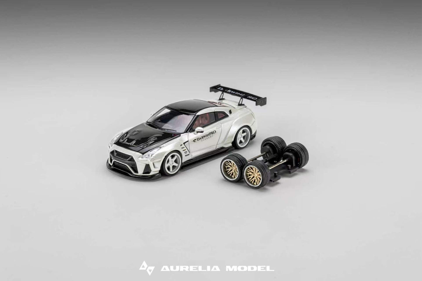 A miniature model of a white and black sports car with decals, shown alongside a matching black chassis with gold-rimmed wheels on a neutral gray gradient background. The brand name 'Aurelia Model' is visible at the bottom.