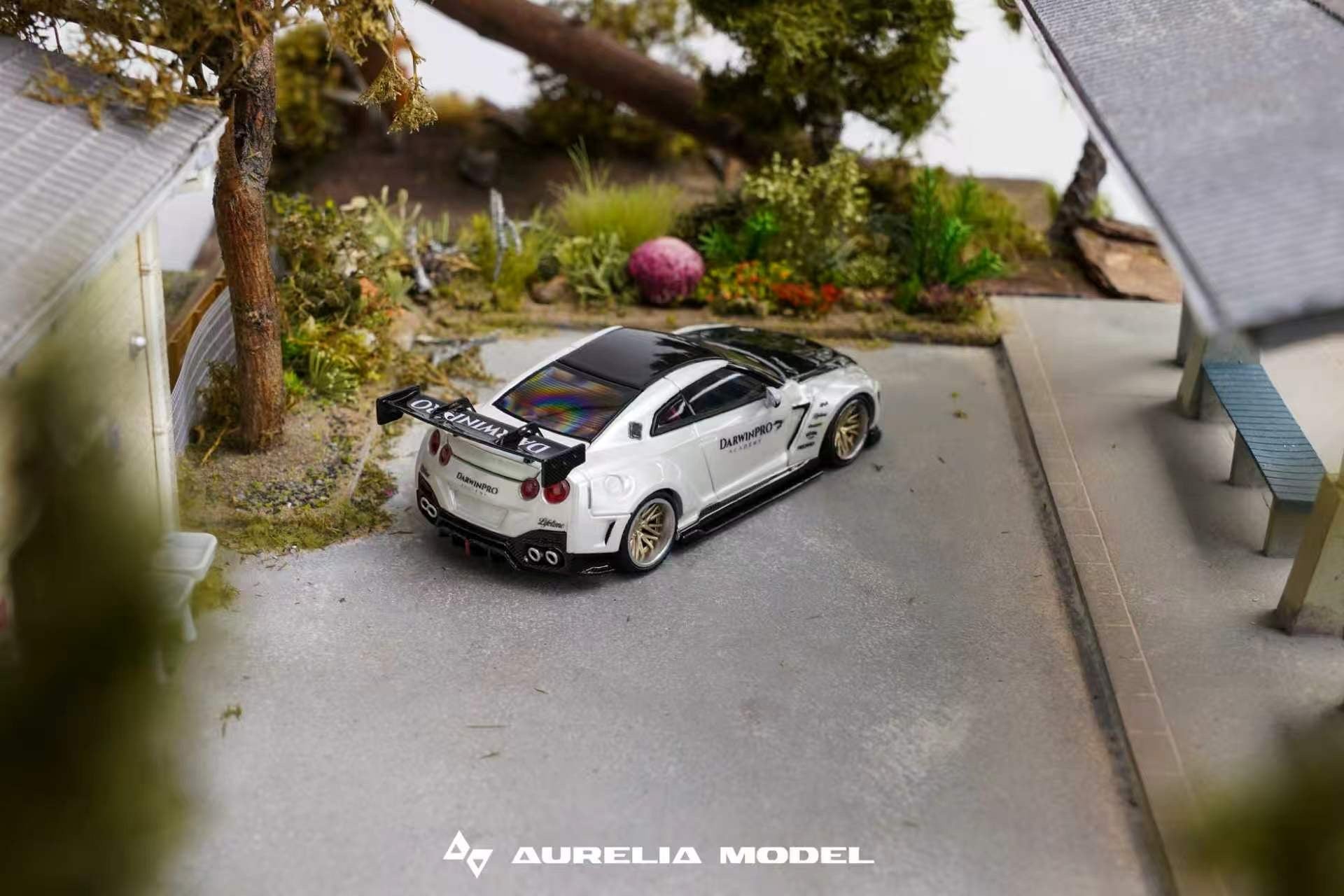 A miniature scale model of a white and black Nissan GT-R Nismo race car, displayed in a detailed diorama setting with a miniature road, trees, and buildings. The brand 'Aurelia Model' is visible as a watermark at the bottom.