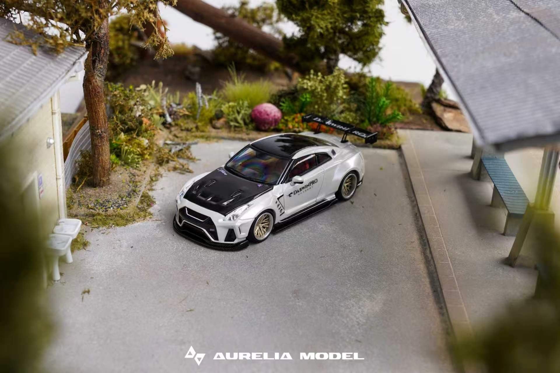 A miniature scale model of a white and black sports car with decals, displayed on a miniature street scene with houses, trees, and a bench. The brand name 'Aurelia Model' is visible at the bottom.