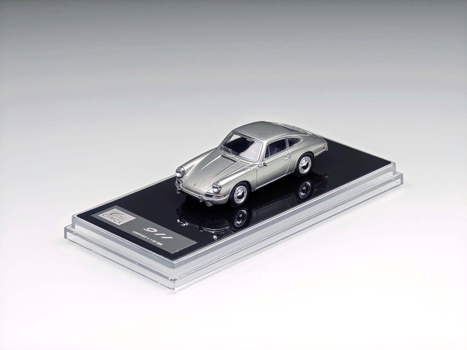 A miniature silver model car displayed on a black rectangular base with a clear acrylic cover, photographed on a clean light gray studio background.