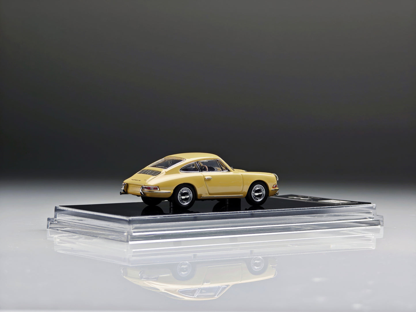A miniature model of a classic yellow Porsche 911 car displayed on a clear acrylic stand with a black base, set against a dark gray studio background.