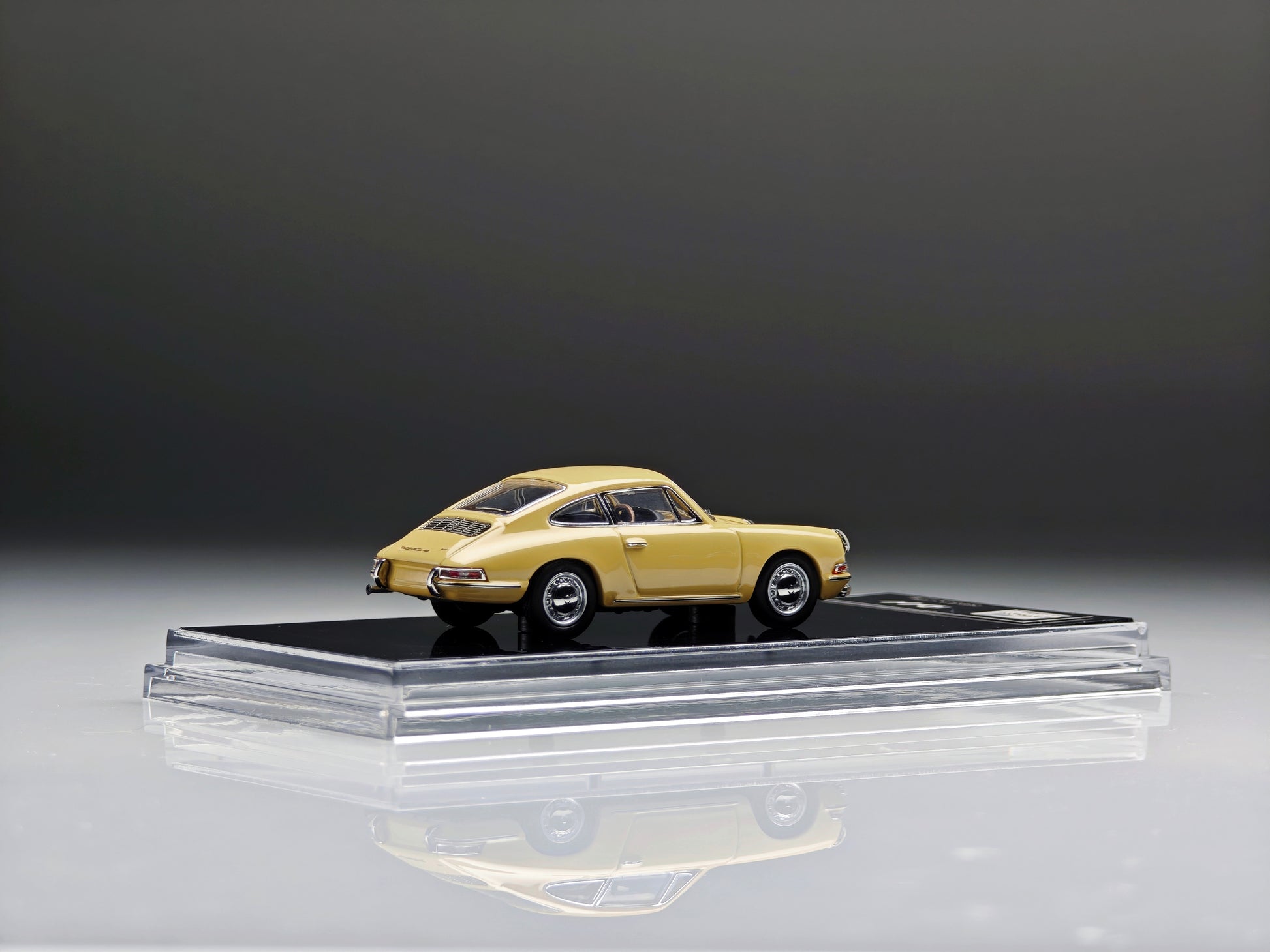 A miniature model of a classic yellow Porsche 911 car displayed on a clear acrylic stand with a black base, set against a dark gray studio background.