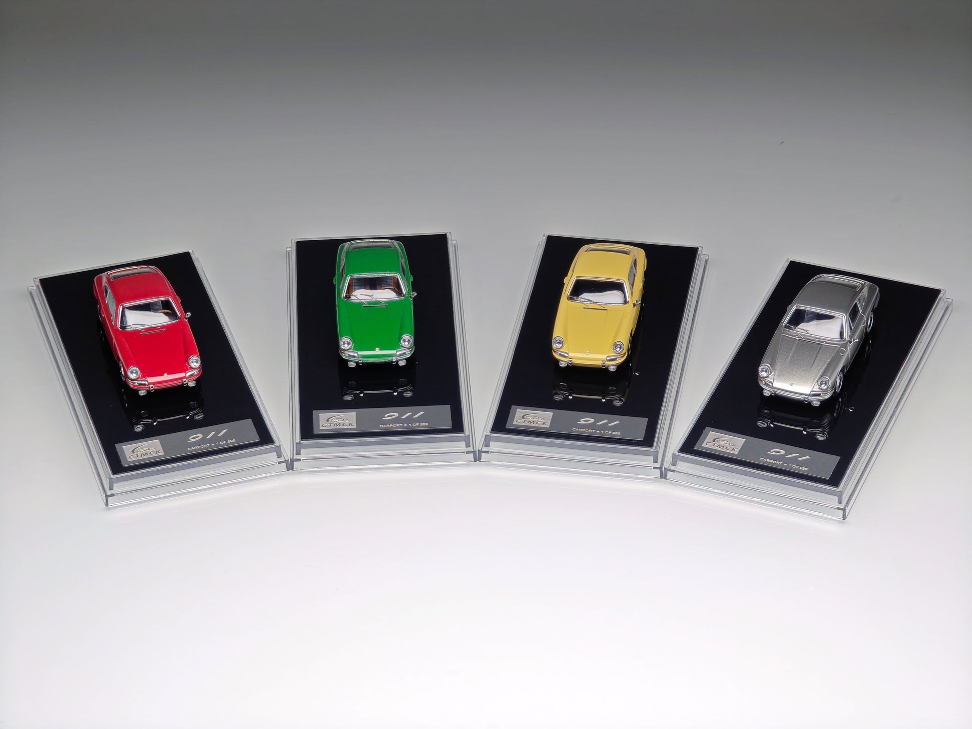 Four miniature model cars in red, green, yellow, and silver, each mounted on a black display card with a clear plastic case, arranged in a row on a light gray background.