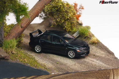 A black diecast model car with a rear spoiler, positioned on a miniature dirt road within a diorama landscape featuring grass, a tree trunk, and a white background. The FiberRun logo is visible in the top right corner.