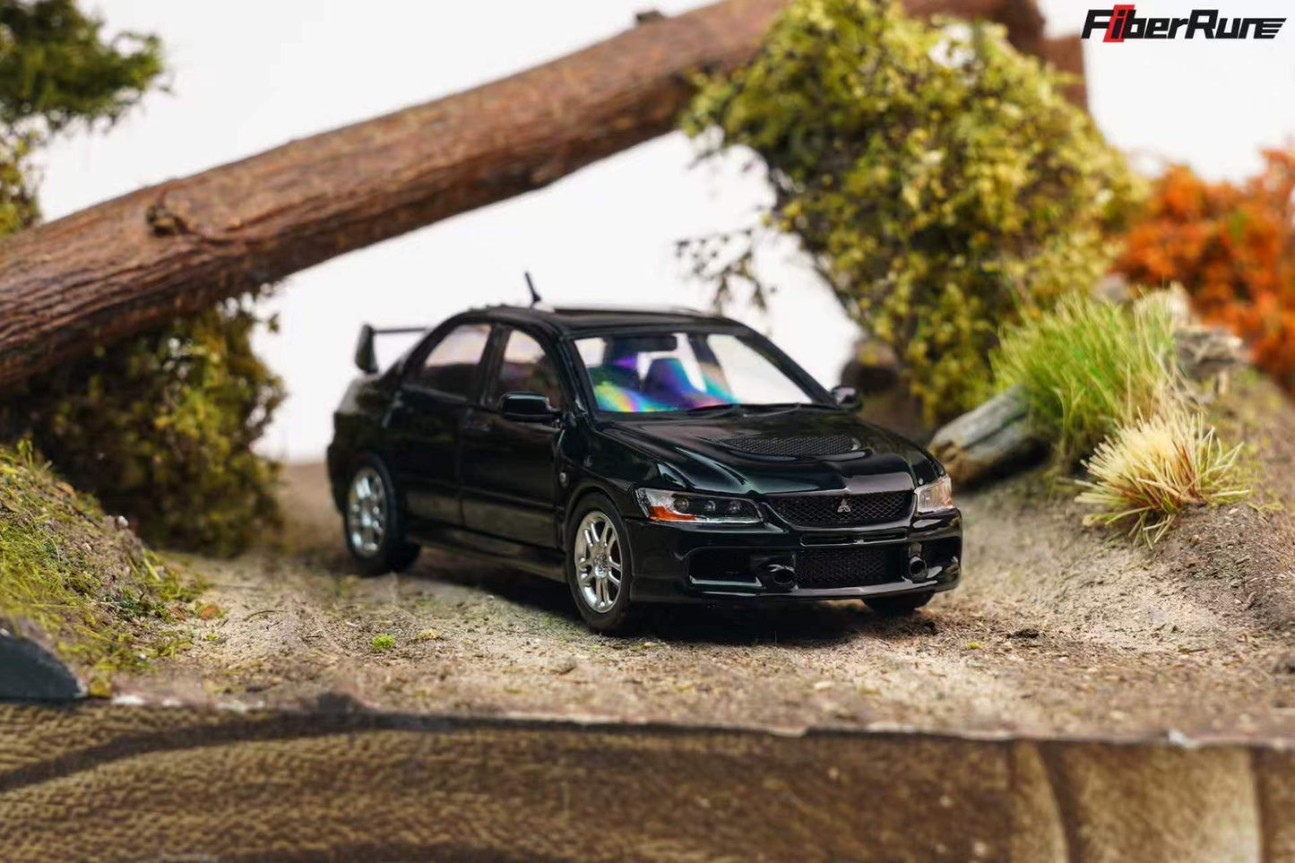 A black diecast model car of a Mitsubishi Lancer Evolution, displayed on a miniature dirt track with grass, moss, and a log backdrop. The FiberRun brand logo is visible in the top right corner.