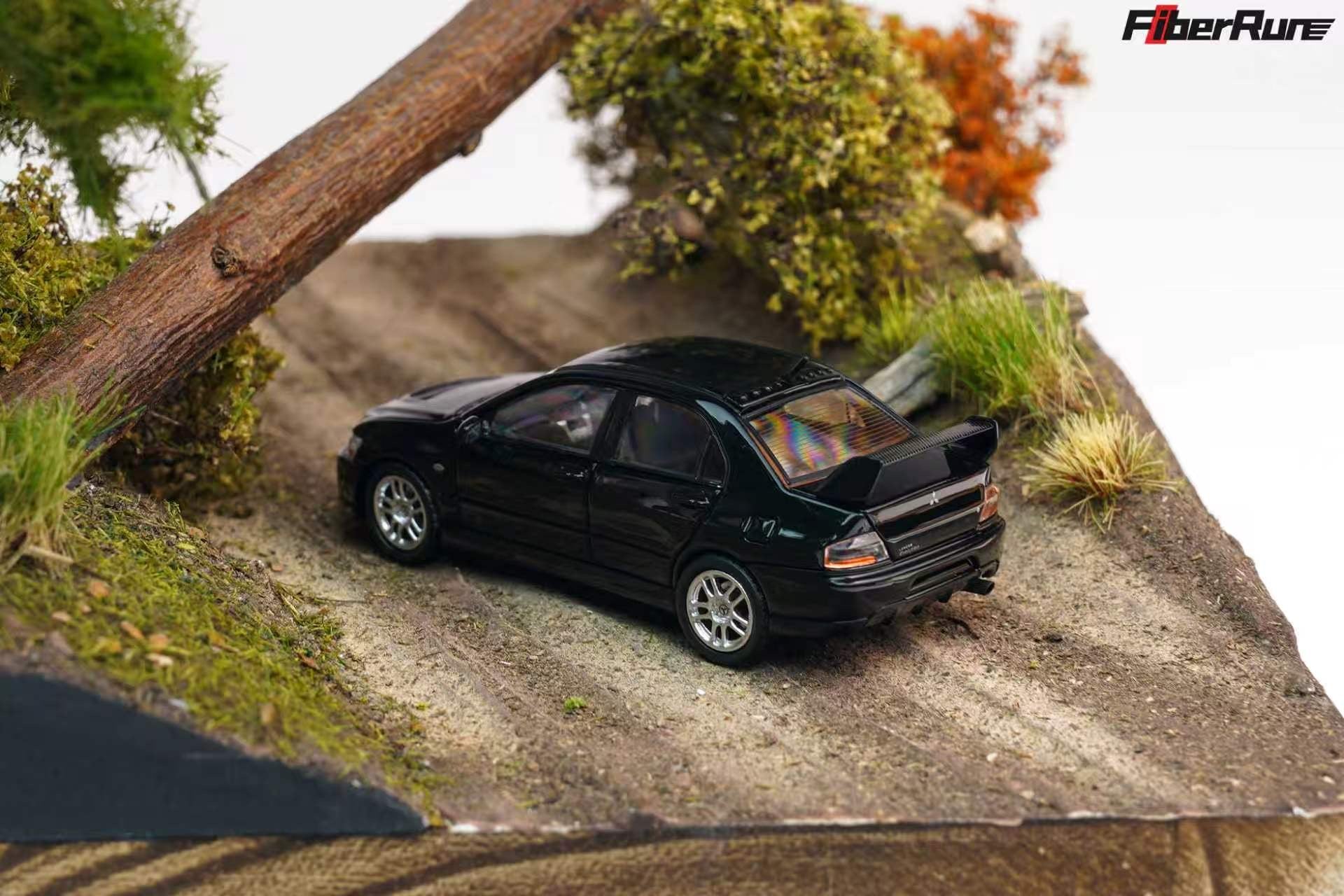 A black diecast model car resembling a Subaru Impreza, positioned on a textured dirt track within a diorama miniature landscape featuring grass, trees, and a large tree trunk.