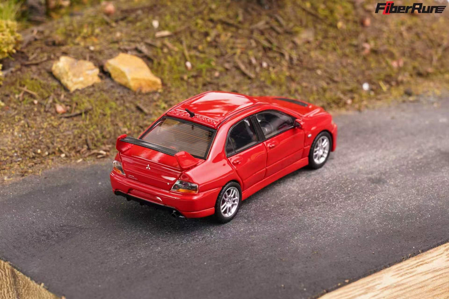A red die-cast model car, specifically a Mitsubishi Lancer Evolution, displayed on a miniature black road within a diorama setup. The background shows a textured brown hillside with small rocks.