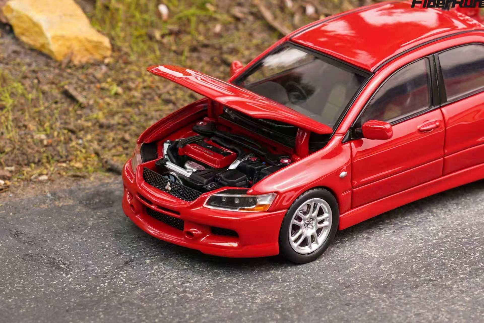 A red die-cast model car with its hood open, showing a detailed engine bay, set on a gray asphalt surface with a blurred outdoor background.