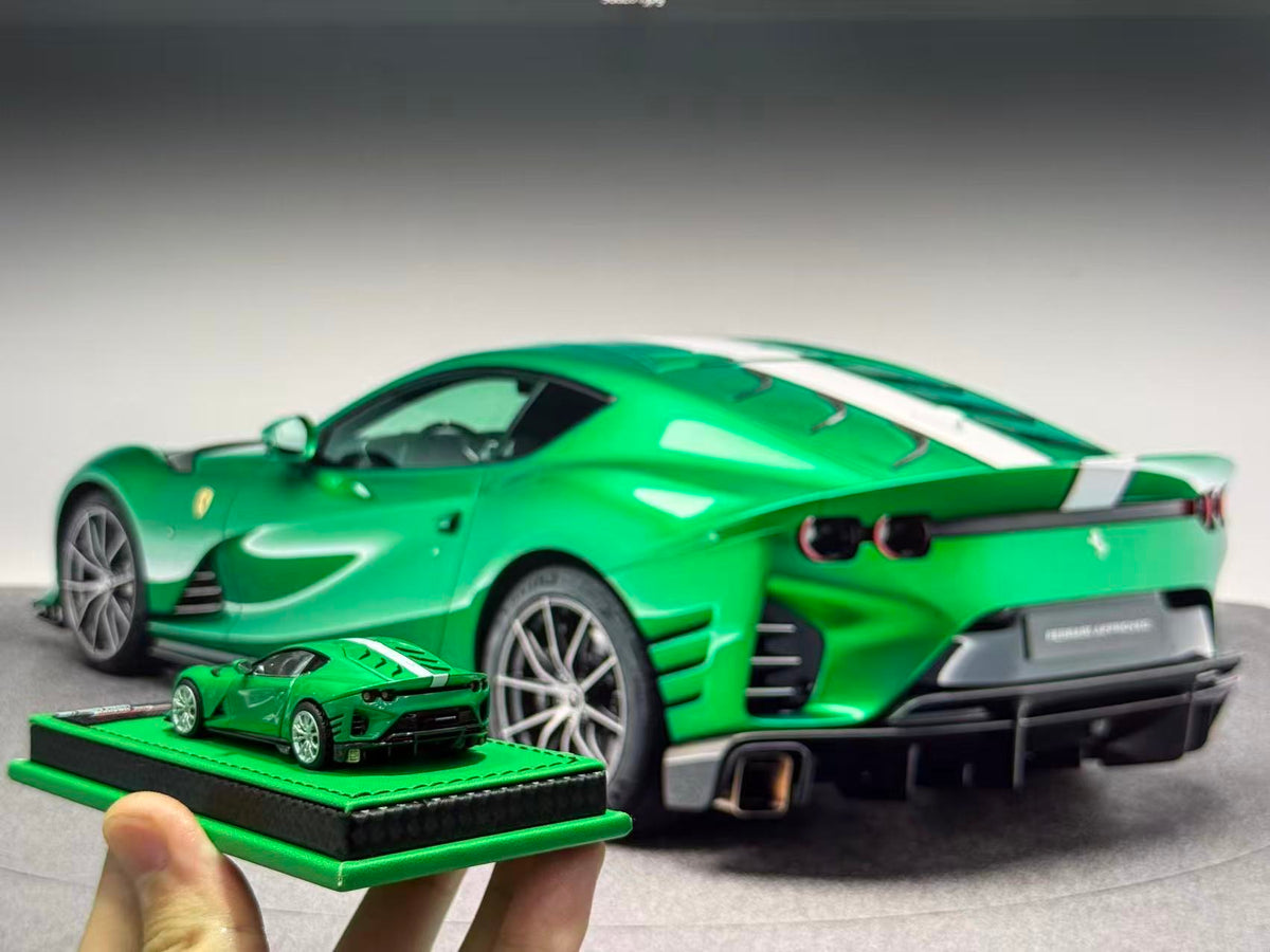 A green die-cast model car displayed on a small green base, held in a hand in the foreground, with a larger matching green sports car blurred in the background.