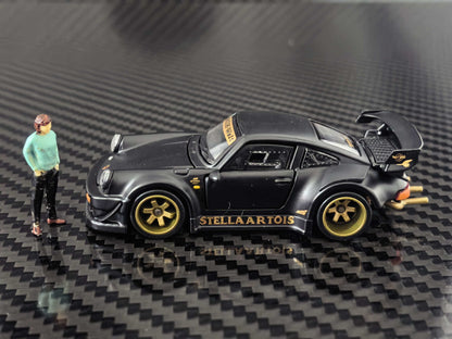 HG Model 1:64 RWB 930 Stella Artois Matte Black diecast model with gold wheels, widebody kit, large rear wing, gold exhaust pipes and included miniature figure, side profile view.