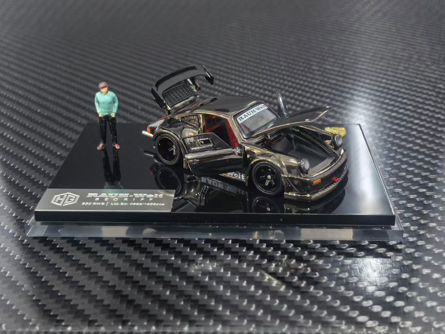 A 1/64 scale model car of a black and gold custom Porsche 911, displayed on a black base with a small figure of a man next to it. The model is photographed on a carbon fiber textured surface.