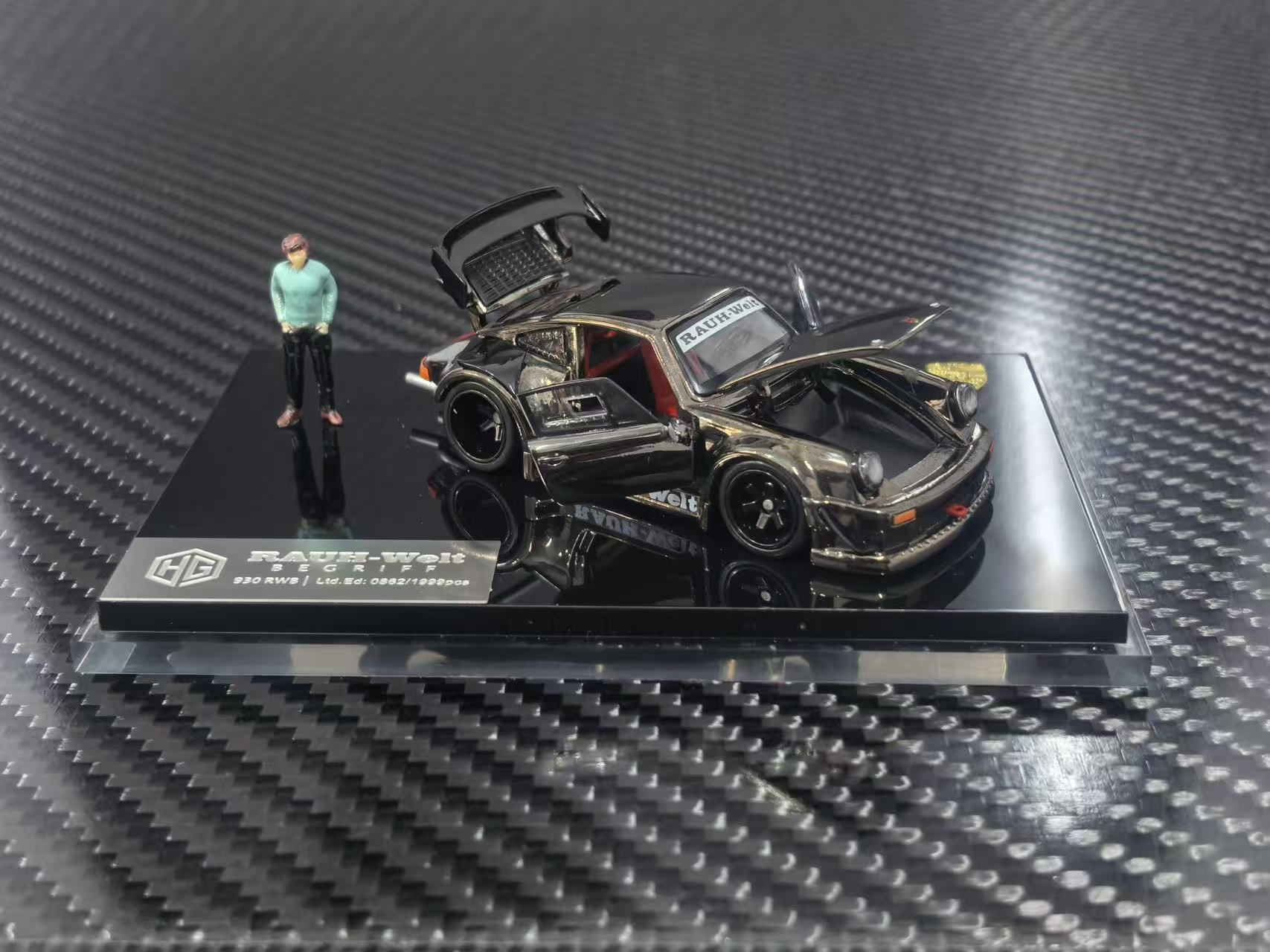 A 1/64 scale model car of a black and gold custom Porsche 911, displayed on a black base with a small figure of a man next to it. The model is photographed on a carbon fiber textured surface.