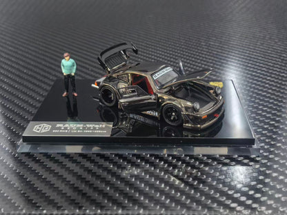 A 1/64 scale model car of a black and gold custom Porsche 911, displayed on a black base with a small figure of a man next to it. The model is photographed on a carbon fiber textured surface.