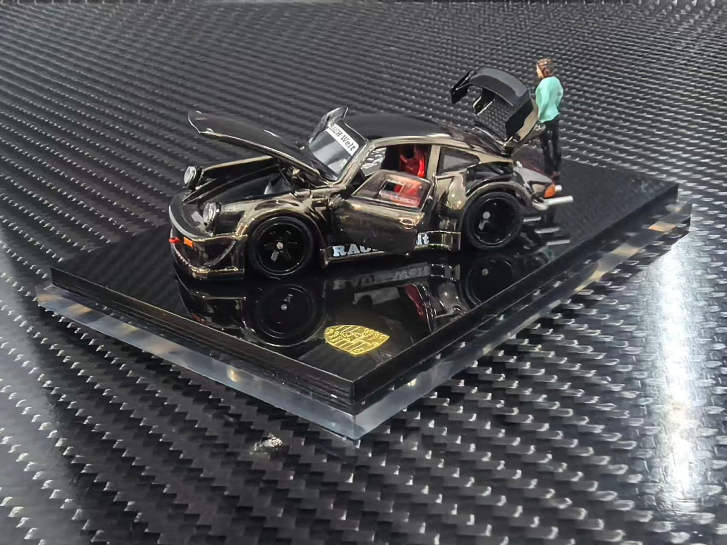 A scale model of a black Porsche 911 race car with its doors open, displayed on a black base with a small miniature figure next to it, set against a carbon fiber textured background.