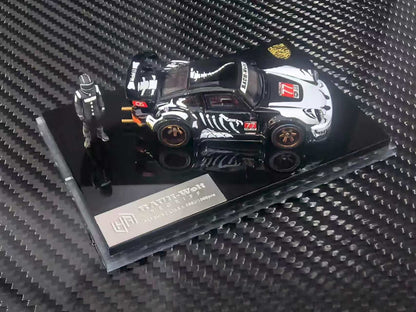 A 1:64 scale diecast model car of a black and white widebody sports car with a small driver figurine, displayed on a black base with a 'Rally-Wagen' plaque, set against a carbon fiber textured background.