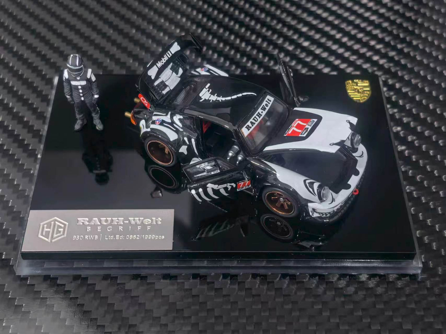 A 1:64 scale model car of a black and white Porsche 911 RSR race car, displayed on a black acrylic base with a small driver figurine. The model is branded by RAUH-Welt and features Porsche branding.