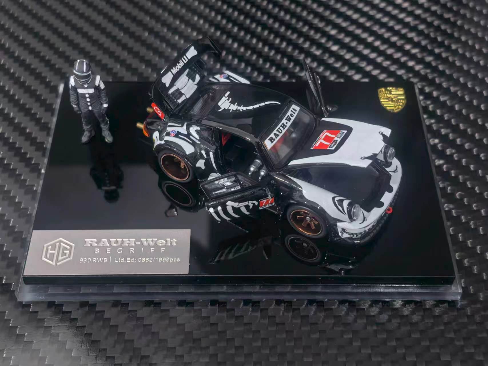 A 1:64 scale model car of a black and white Porsche 911 RSR race car, displayed on a black acrylic base with a small driver figurine. The model is branded by RAUH-Welt and features Porsche branding.