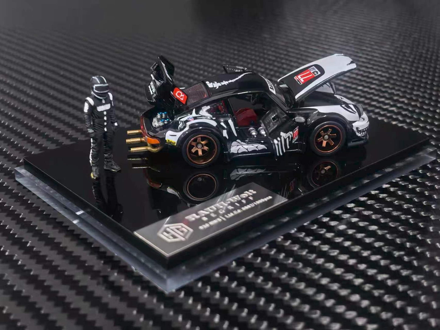 A scale model of a black and white racing Porsche 911 with a small driver figurine, displayed on a black base with a carbon fiber pattern background.