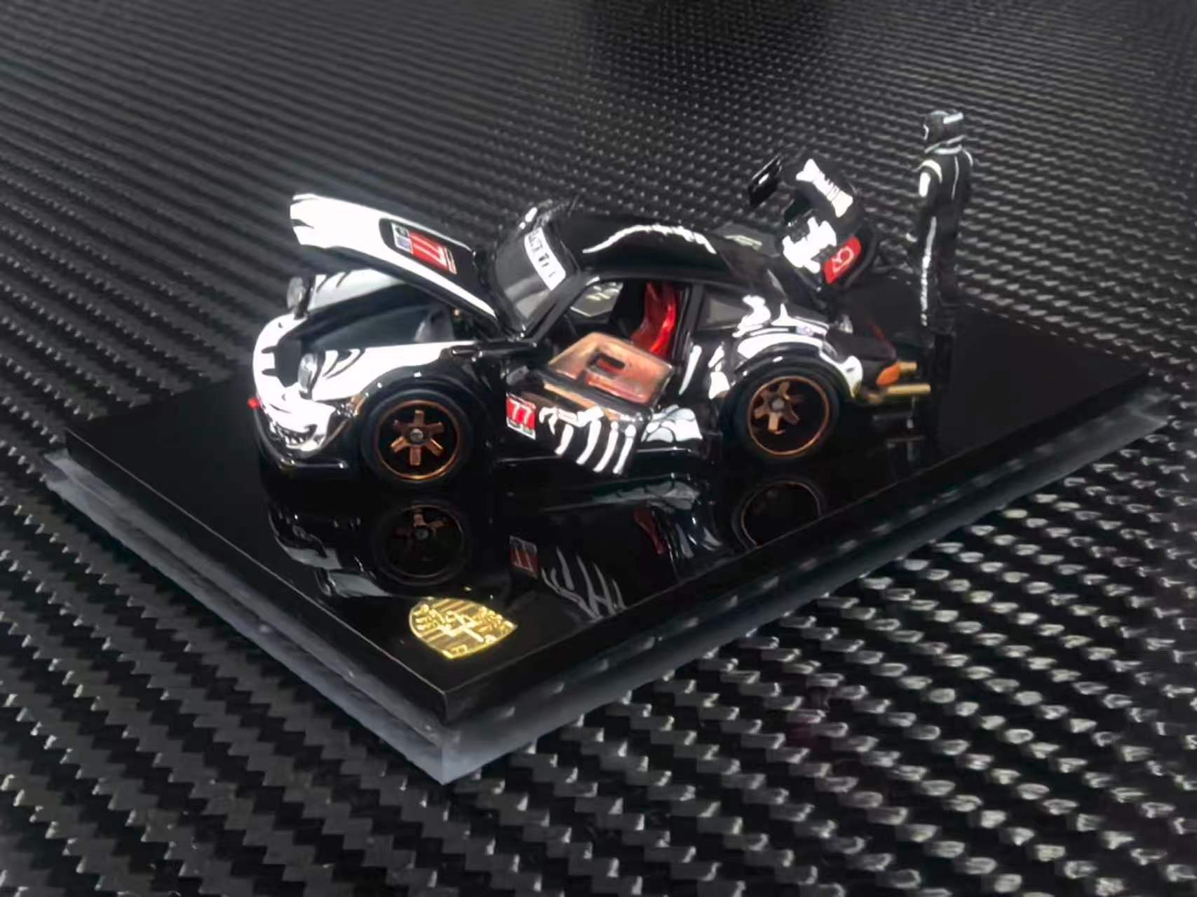 A black and white diecast model Porsche 911 Carrera RSR with orange wheels, displayed on a black base with a gold crest, set against a carbon fiber textured background.