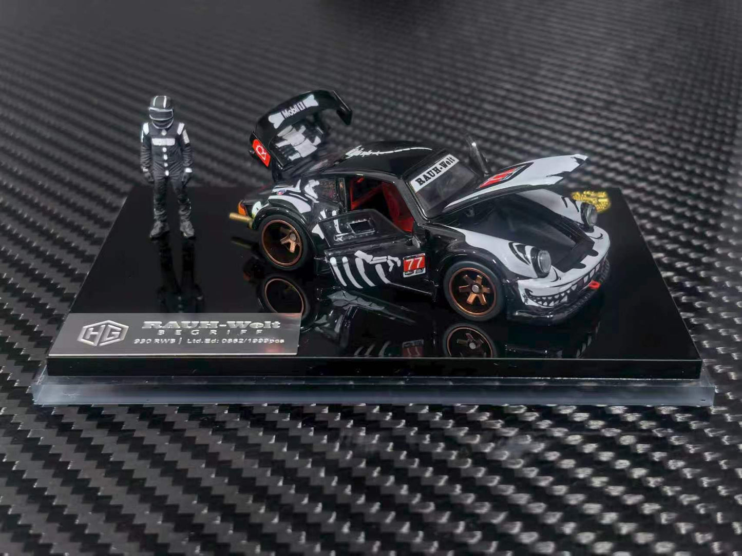 A 1/64 scale diecast model car of a black and white K100 drift car with a driver figure, displayed on a black base over a carbon fiber textured background.