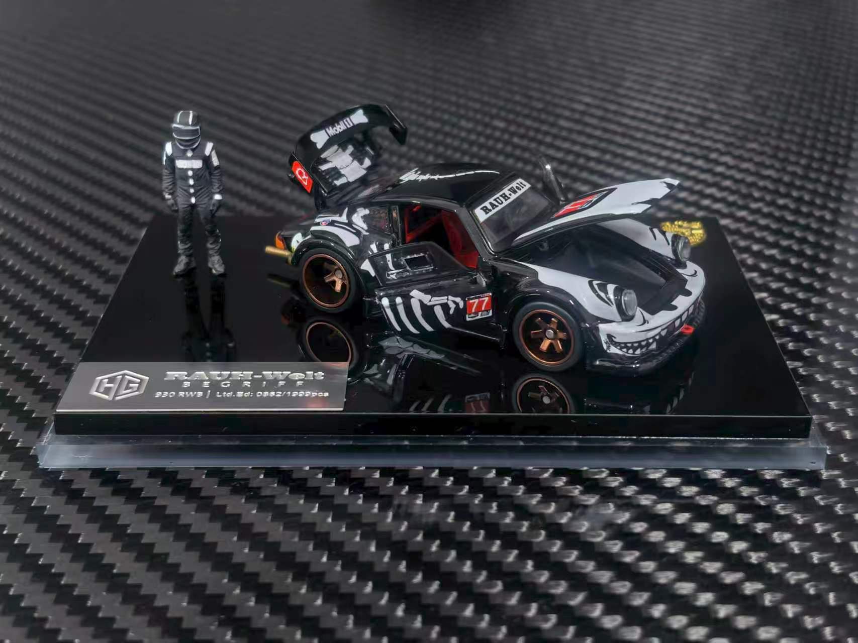 A 1/64 scale diecast model car of a black and white K100 drift car with a driver figure, displayed on a black base over a carbon fiber textured background.