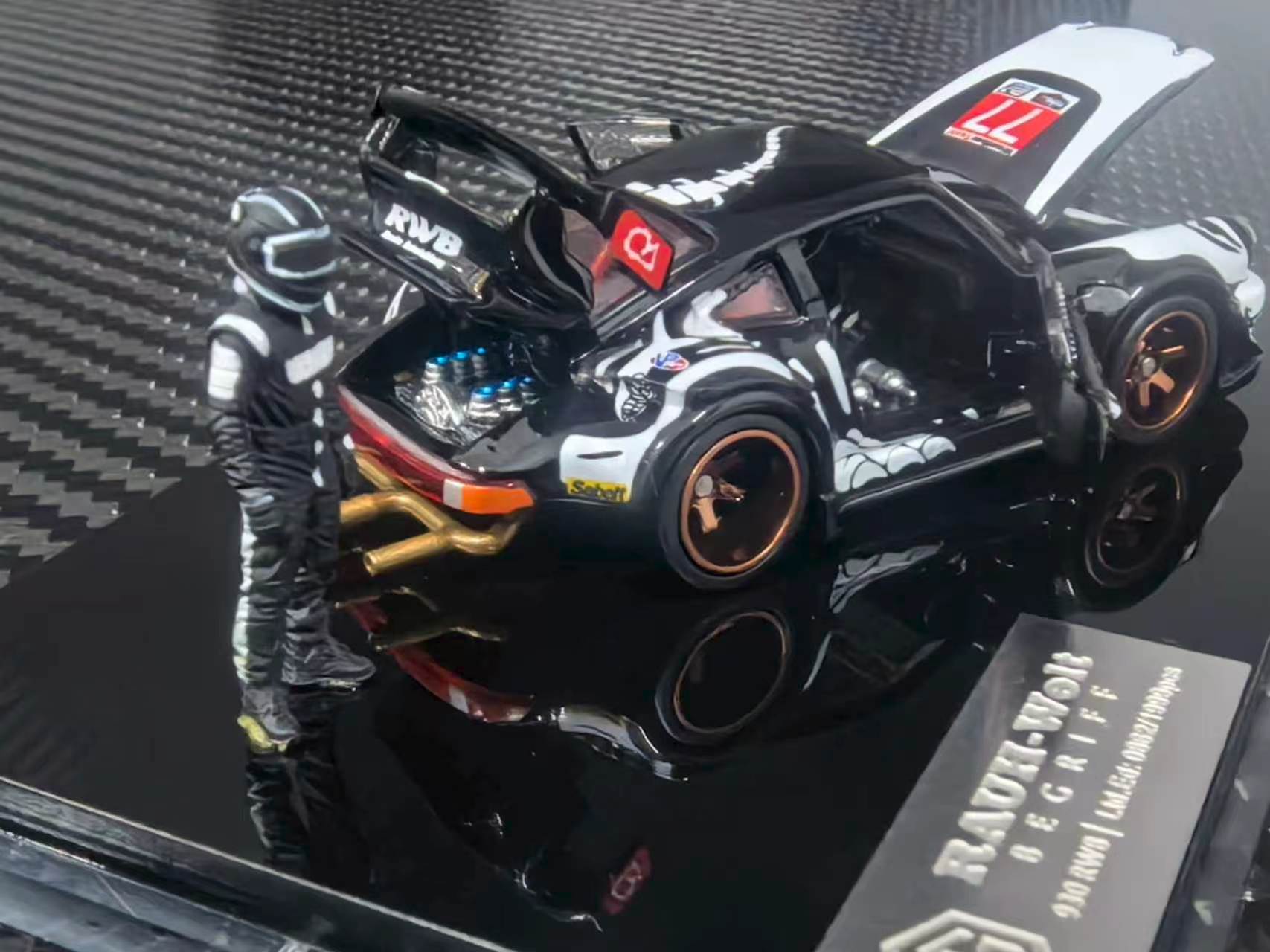 A 1:64 scale model car of a black and white Porsche 911 with custom racing livery, shown with its doors open next to a small driver figurine on a glossy black display base.