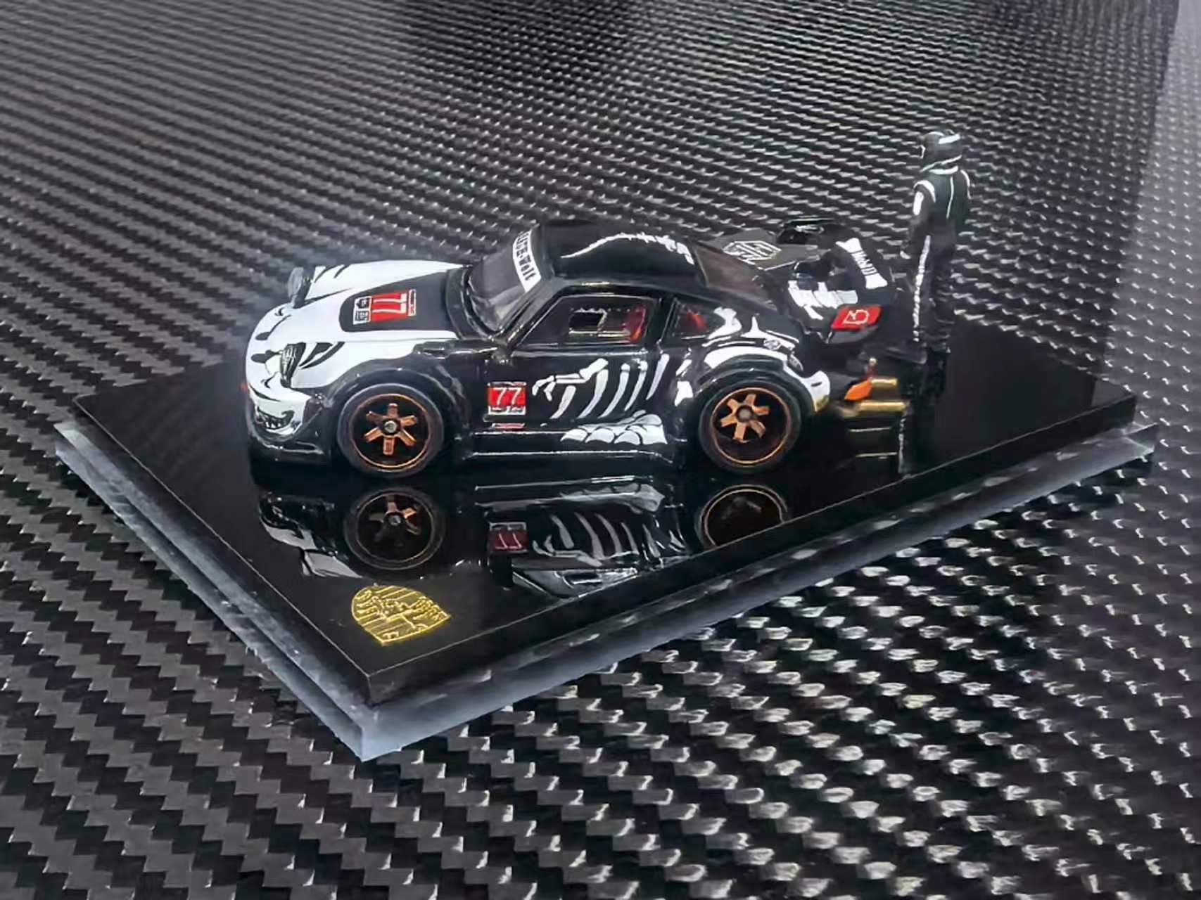 A scale model of a black and white drift racing car with orange wheels, displayed on a black acrylic base over a carbon fiber patterned surface.