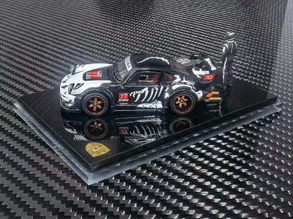 A scale model of a black and white drift racing car with orange wheels, displayed on a black acrylic base over a carbon fiber patterned surface.