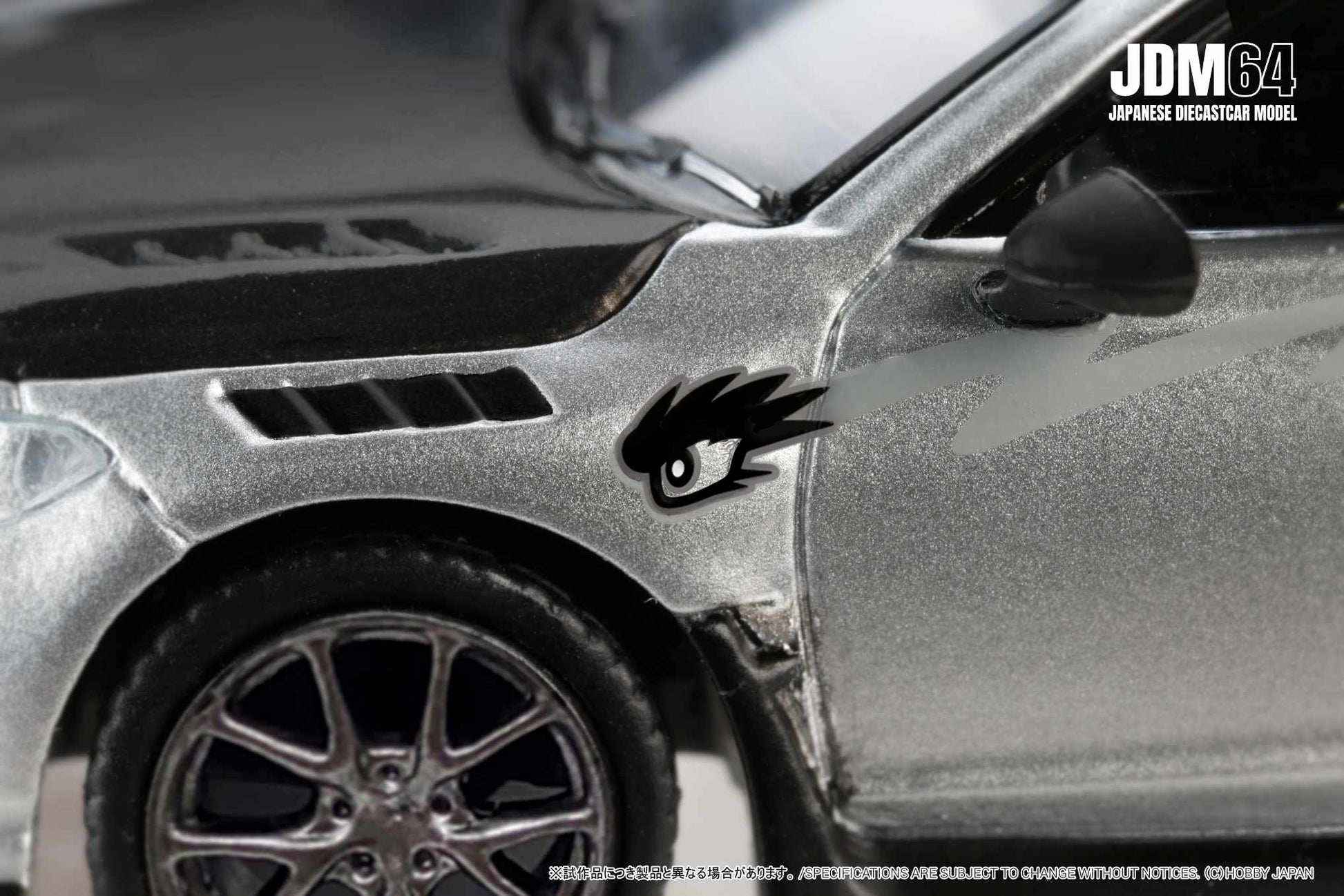 Close-up of a silver diecast car model with black details and stylized eye decal. The text "JDM64 Japanese Diecast Car Model" is in the corner.